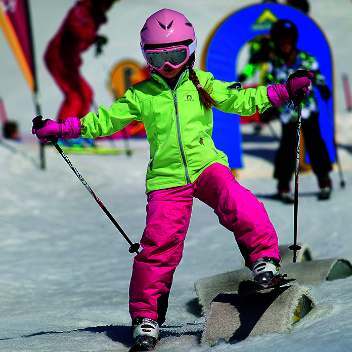 Kinderskilauf Geländeform Kind in pink-grünem Ski-Anzug und Skiern über Hindernissen